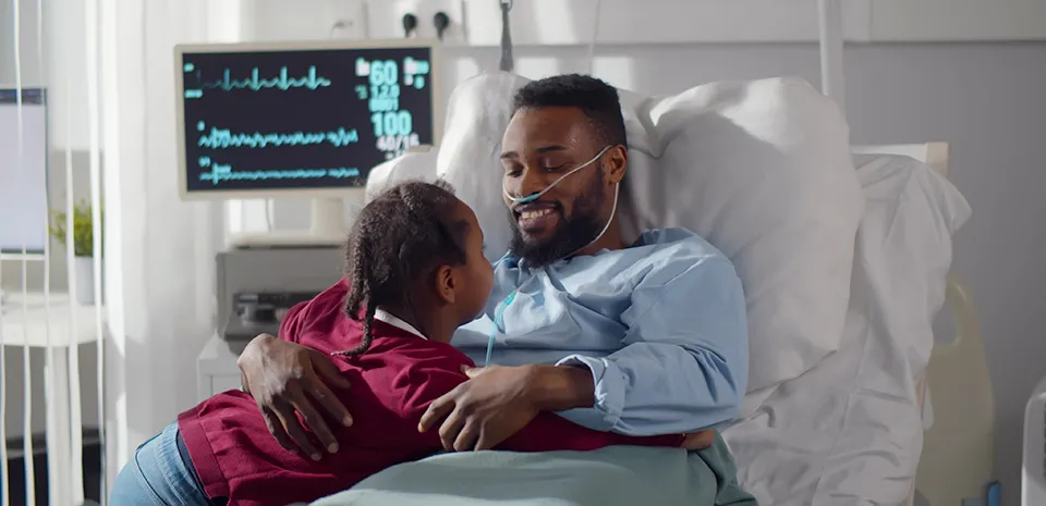 A daughter visits her father who is in a hospital bed with his vitals on a monitor behind him.