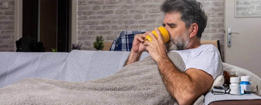 A man is under a blanked on a couch and drinking from a mug.