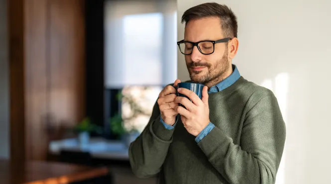 Man with eyes closed holding a warm cup of coffee in a kitchen practicing mindfulness